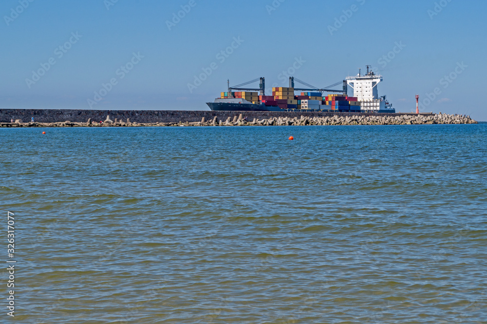 A container ship loaded with metal containers of different colors enters the coastal bay past a gray stone pier with a red and white lighthouse against the blue sky and the coastal surface of the sea