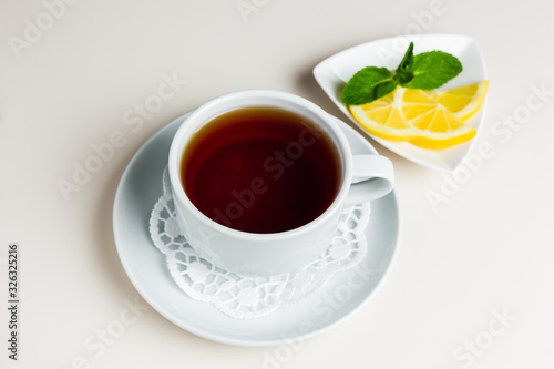 A white cup of black tea on a saucer and paper napkin, a small saucer with sliced lemon and mint