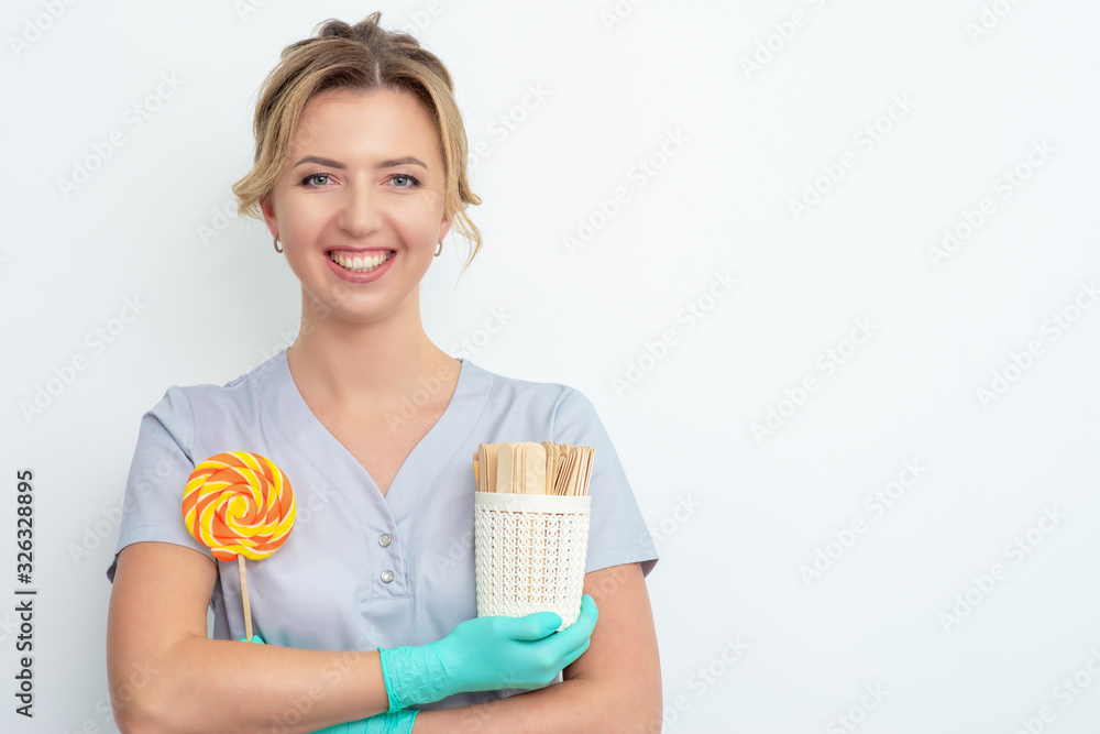 Smiling beautician holds lollipop and sticks for waxing epilation or sugaring on white background, copy space. Smiling beautician holds lollipop and sticks for waxing epilation or sugaring on white background, copy space.