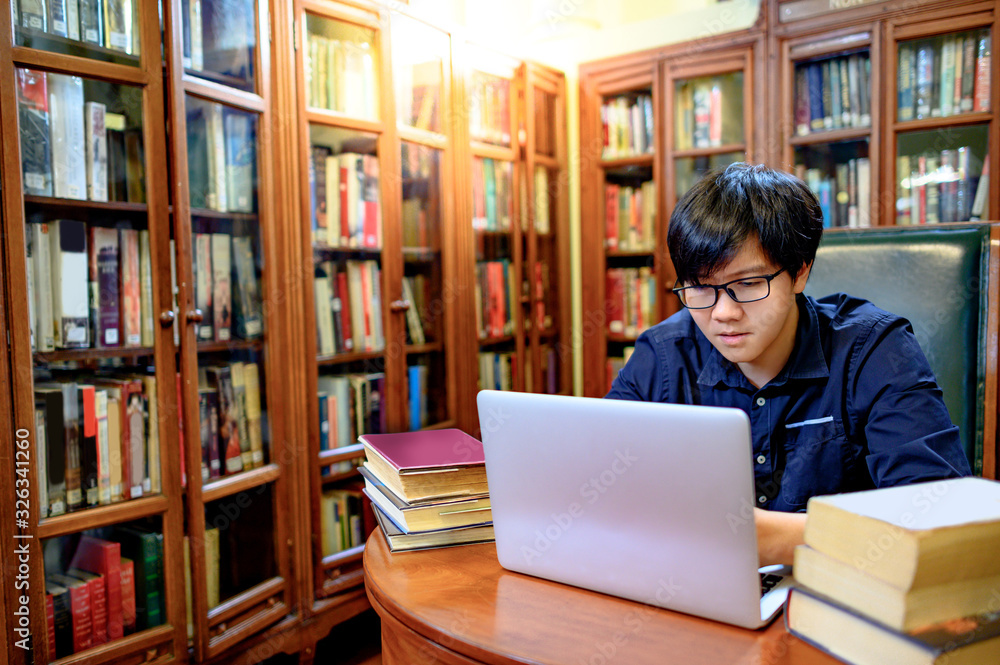 Asian man university student using laptop computer and reading book nearby vintage bookcase or bookshelf in college library. Textbook resources for education research. Scholarship opportunity concept Asian man university student using laptop computer and reading book nearby vintage bookcase or bookshelf in college library. Textbook resources for education research. Scholarship opportunity concept
