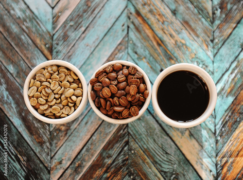 Green raw beans, roasted coffee beans and black coffee in three white cups on turquoise wooden background top view