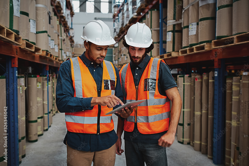 © StratfordProductions - Young focused workers in warehouse discussing work on digital tablet