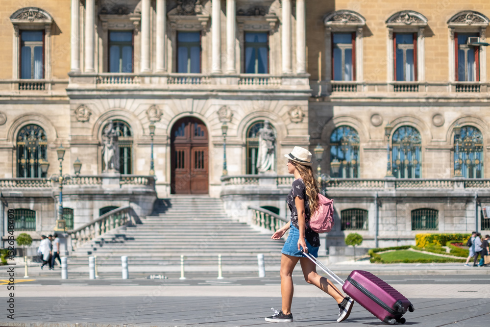 A woman tourist walks by in the city carying her suitcase and a hat.