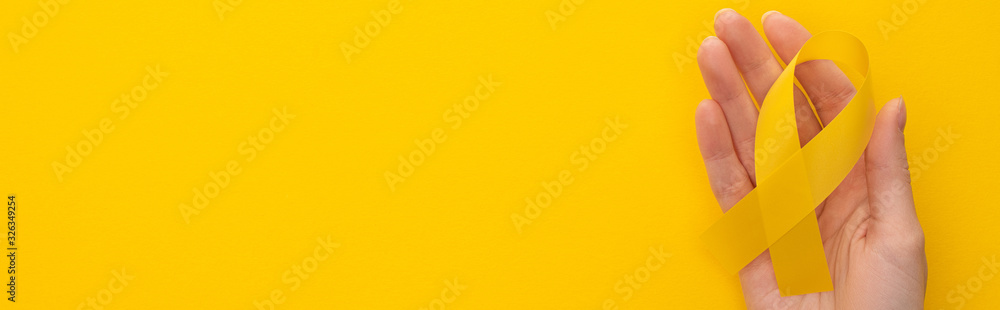 Partial view of female hand with yellow ribbon on colorful background, panoramic shot, international childhood cancer day concept Partial view of female hand with yellow ribbon on colorful background, panoramic shot, international childhood cancer day concept