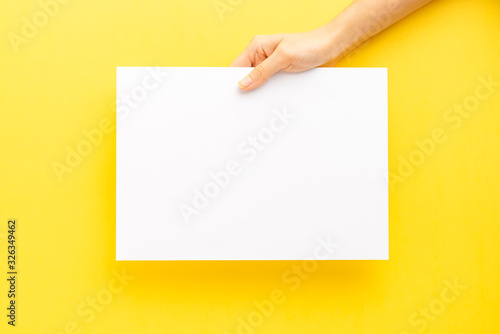 Female hand holding empty paper sheet on yellow background. Top view