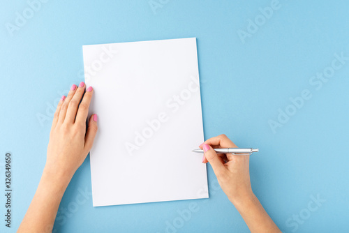 Overhead shot of female hands writing with pen over empty white sheet of paper on blue background photo