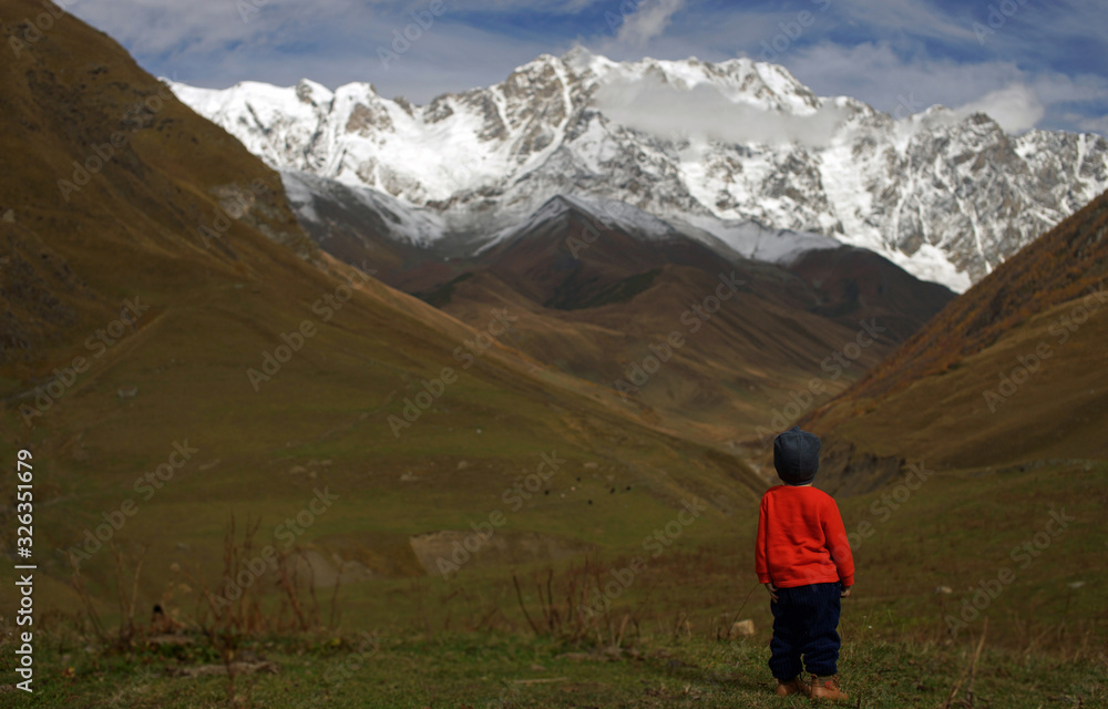 Little boy standing in front of snowy mountaines and autumn valley. Small traveller in wild nature, view from behind