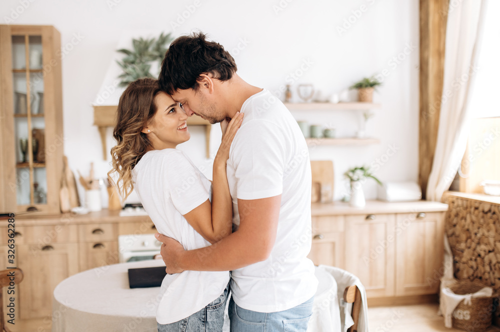 The hugging and smiling happy couple in white t-shirts at home in the kitchen The hugging and smiling happy couple in white t-shirts at home in the kitchen