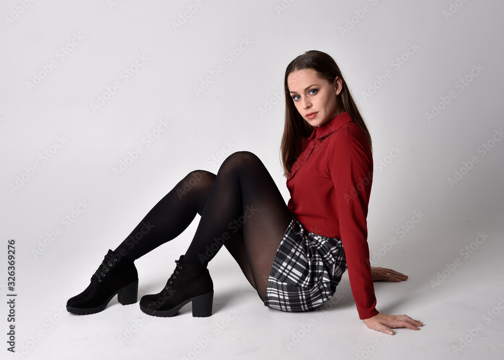 full length portrait of a pretty brunette girl wearing a red shirt and  plaid skirt with leggings and boots. Seated pose on a studio background.  Stock Photo