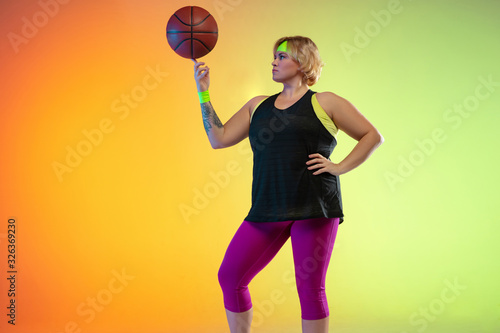 Young caucasian plus size female model's training on gradient orange background in neon light. Doing workout exercises with the ball. Concept of sport, healthy lifestyle, body positive, equality. © master1305
