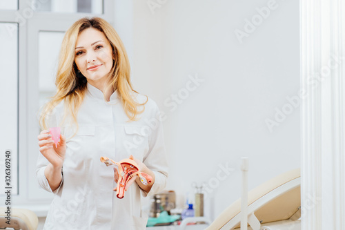 Doctor gynecologist holding anatomical model of uterus and menstrual cup, talking about intimate hygiene menstrual eco cup, how use, to female patient in an chair in modern medical office. photo