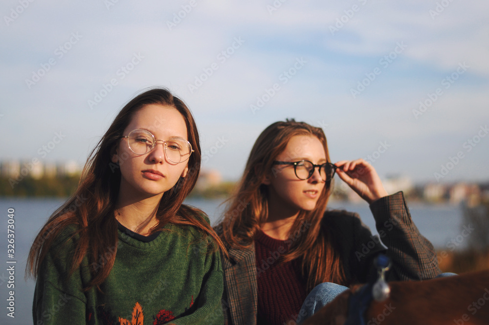 Two fashionable girls in sunglasses , girlfriends, are sitting on the pier near the river, the beach.Two girlfriends with model appearance.Sunset time.