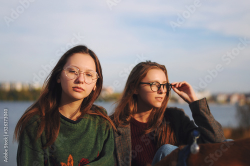 Two fashionable girls in sunglasses , girlfriends, are sitting on the pier near the river, the beach.Two girlfriends with model appearance.Sunset time.