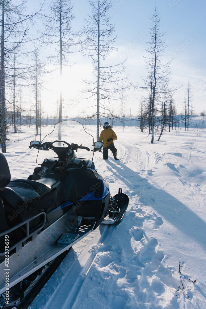 a young guy in a yellow warm jacket stands near his snowmobile in the forest on very deep snow on a frosty winter day