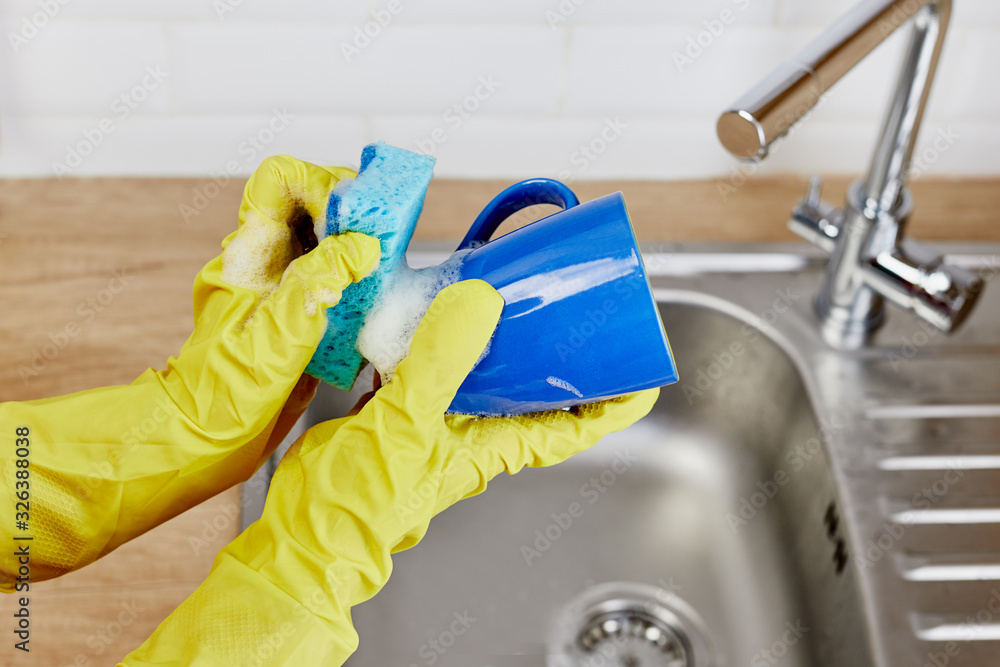 Hands with sponge wash the cup under water, housewife woman in yellow rubber protective gloves washing blue mug