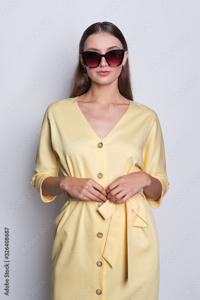 Fashion model in big sunglasses wearing yellow dress with buttons posing over gray background Fashion model in big sunglasses wearing yellow dress with buttons posing over gray background