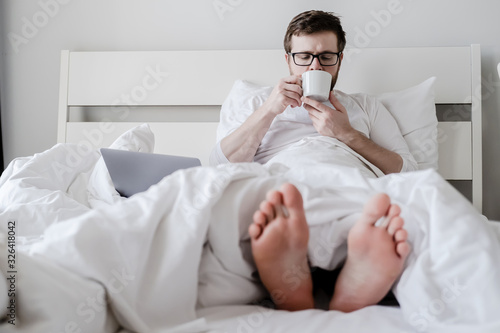 Relaxed man, in pajamas, drinks aromatic coffee, sits in bed with his feet out from under the covers, a laptop lies nearby.