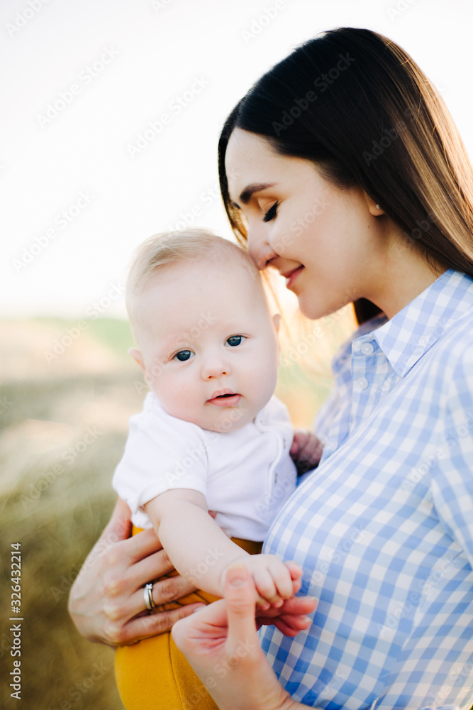 a young happy mother with a small child in her arms, gently leaning against his head, stands in the middle of a field near a collected haystack in the rays of the sunset, summer sun a young happy mother with a small child in her arms, gently leaning against his head, stands in the middle of a field near a collected haystack in the rays of the sunset, summer sun