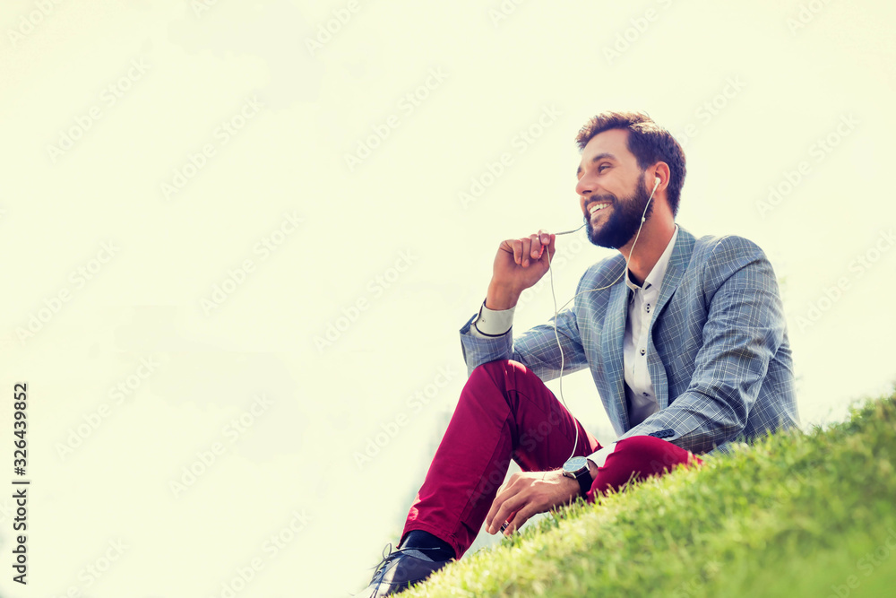 Young attractive businessman talking on smartphone with earphones on while sitting on the grass Young attractive businessman talking on smartphone with earphones on while sitting on the grass