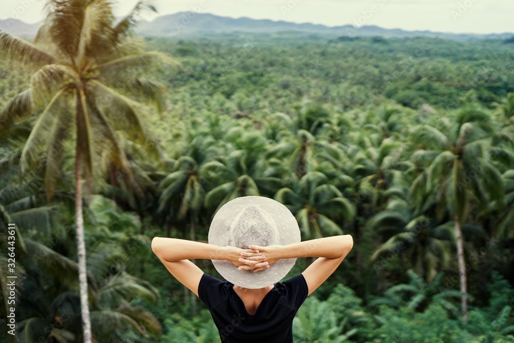Fototapeta premium Tropical vacation. Young woman in hat enjoying the view of beautiful coconut palms plantation.