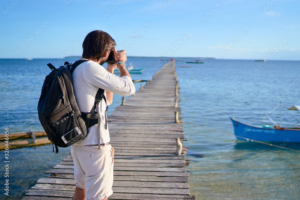 Photography and travel. Young man with rucksack standing on wooden fishing pier taking photo of beautiful tropical sea view.