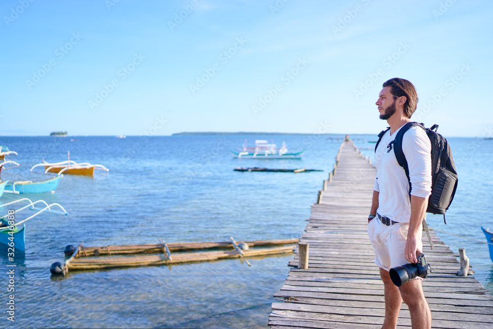 Photography and travel. Young man with rucksack holding camera standing on wooden fishing pier enjoying beautiful tropical sea view.