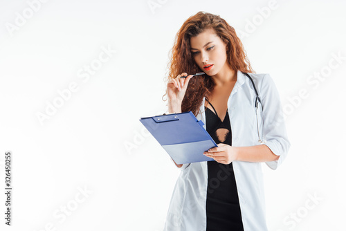 curly and sexy nurse holding pen and looking at clipboard isolated on white