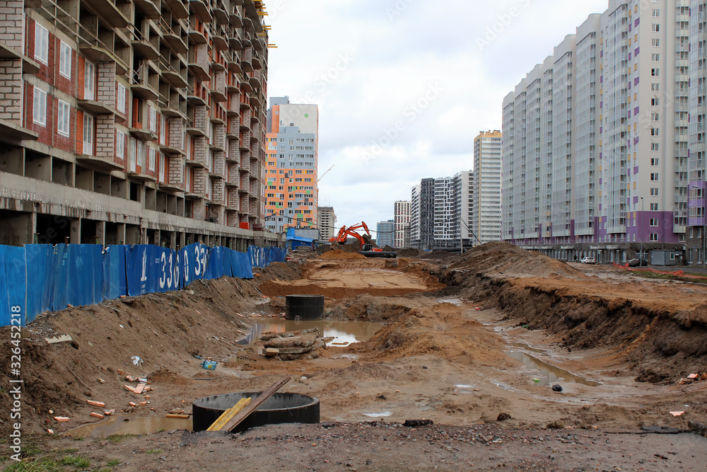 Road. Mountains of sand. Two excavators dig a hole. Around you can see multi-story buildings.