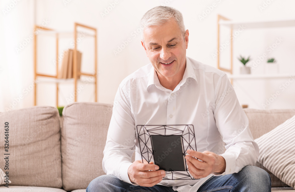 Cheerful aged man sitting on couch holding photo frame Cheerful aged man sitting on couch holding photo frame