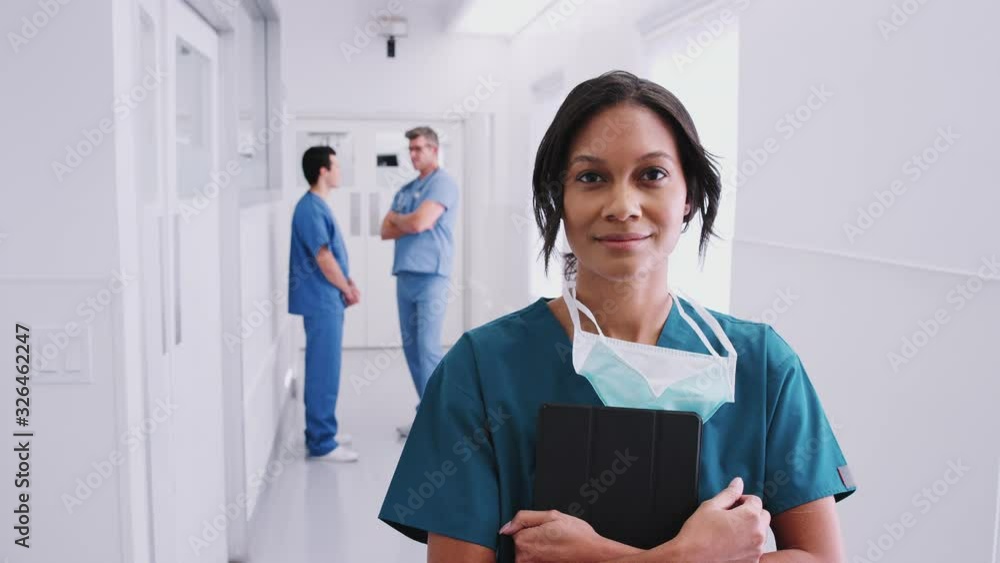Female Surgeon Wearing Scrubs And Mask In Busy Hospital Corridor With Colleagues In Background Female Surgeon Wearing Scrubs And Mask In Busy Hospital Corridor With Colleagues In Background