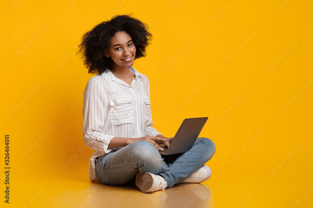 Afro woman sitting on floor, using laptop and smiling at camera Afro woman sitting on floor, using laptop and smiling at camera