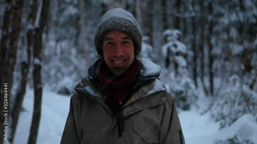Fixed angle of a man standing and smiling at the camera while snow falls on his head and shoulders in the wintertime, with a forest background Fixed angle of a man standing and smiling at the camera while snow falls on his head and shoulders in the wintertime, with a forest background