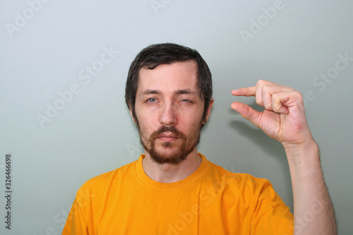 Middle-aged man with a mustache and a brunette beard in a yellow T-shirt on a gray background with a narrowed eye, he shows with his fingers something small