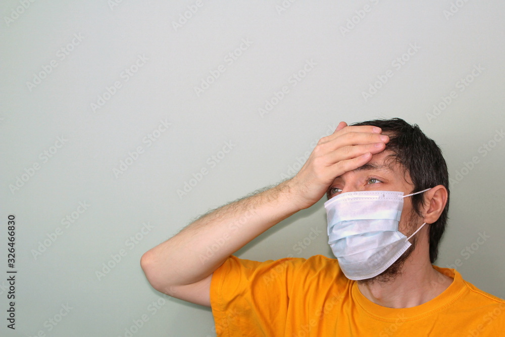 Middle-aged adult man in a yellow T-shirt on a gray background in a medical mask holds his hand near his forehead. Headache, high fever. Disease protection coronavirus epidemic problem from China Middle-aged adult man in a yellow T-shirt on a gray background in a medical mask holds his hand near his forehead. Headache, high fever. Disease protection coronavirus epidemic problem from China