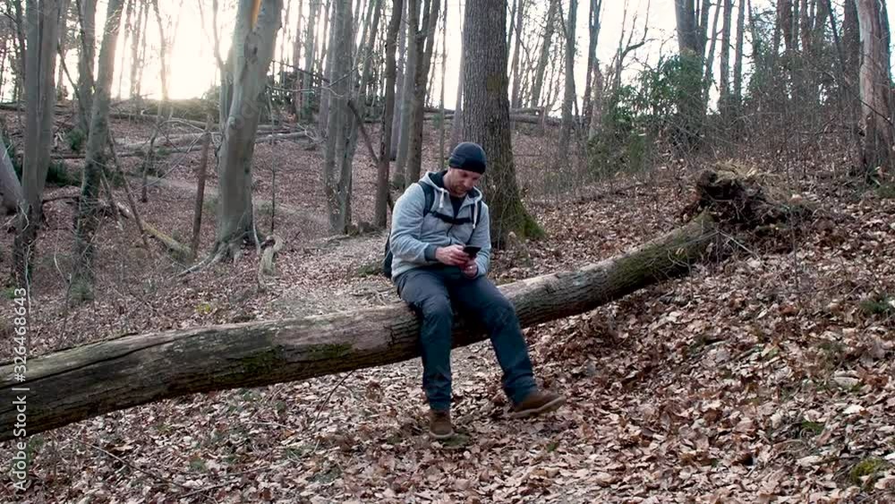 Hiker texting on his smartphone while sitting on a broken tree in a forest. Hiker texting on his smartphone while sitting on a broken tree in a forest.
