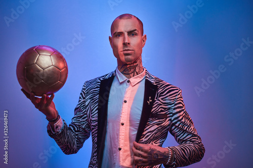 Plakat Fashionable, handsome, tattooed, bald male model posing in a studio for the photoshoot wearing fashionable custom made zebra striped style tuxedo and rose patterned shirt, holding a golden soccer ball