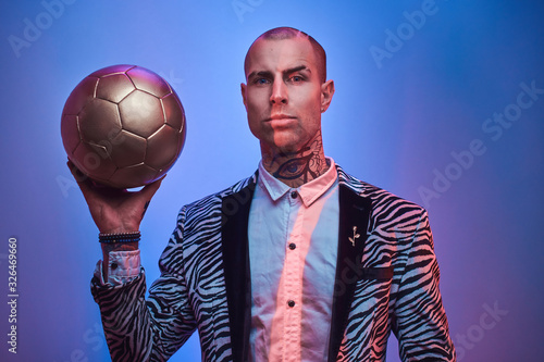 Plakat Fashionable, handsome, tattooed, bald male model posing in a studio for the photoshoot wearing fashionable custom made zebra striped style tuxedo and rose patterned shirt, holding a golden soccer ball