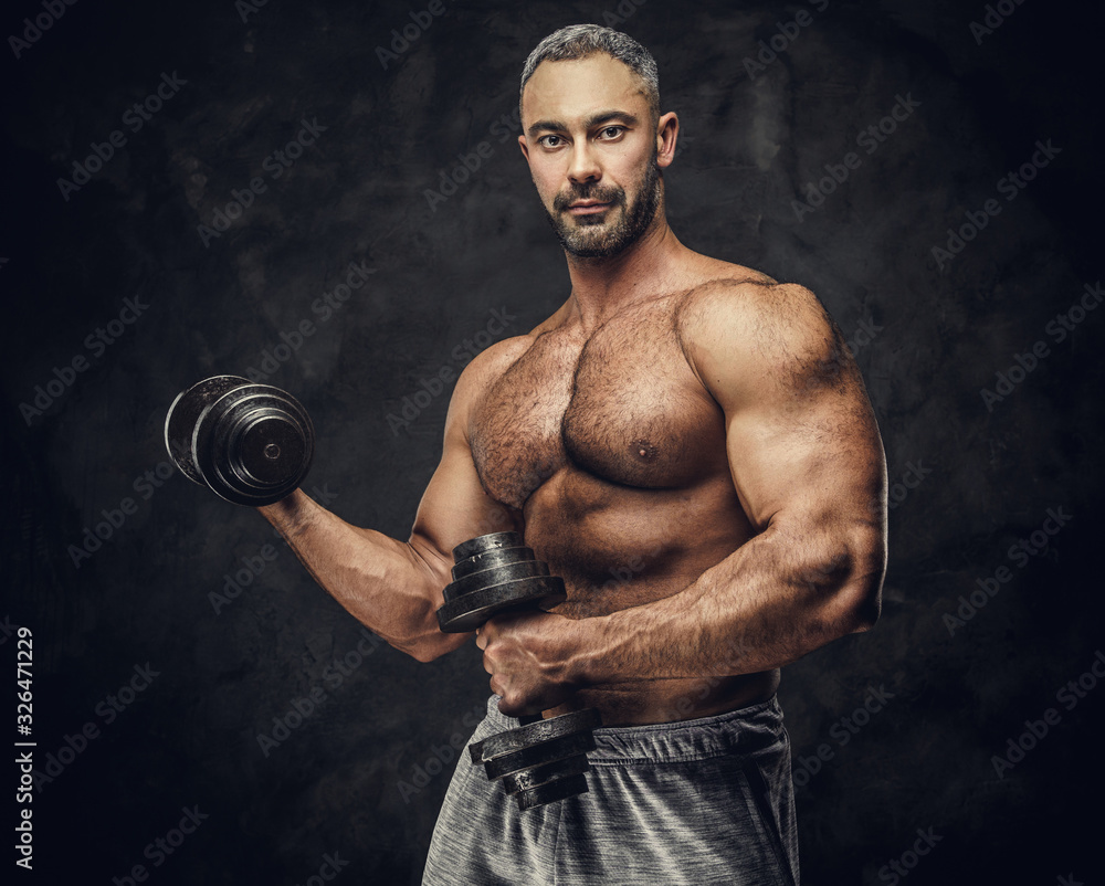 Strong, adult, fit muscular caucasian man coach posing for a photoshoot in a dark studio under the spotlight wearing grey sportswear, showing his muscles and putting up a dumbbells looking confident