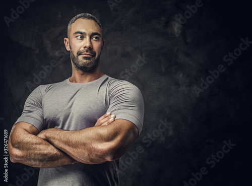 Strong, adult, fit muscular caucasian man posing for a photoshoot in a dark studio under the spotlight wearing grey sportswear, showing his muscles with arms crossed, looking confident and calm photo