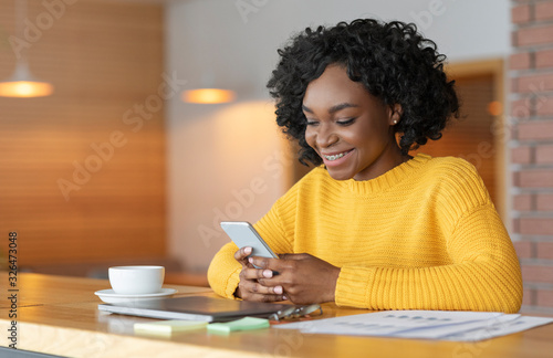 Cheerful afro girl chatting at cafe, using smartphone