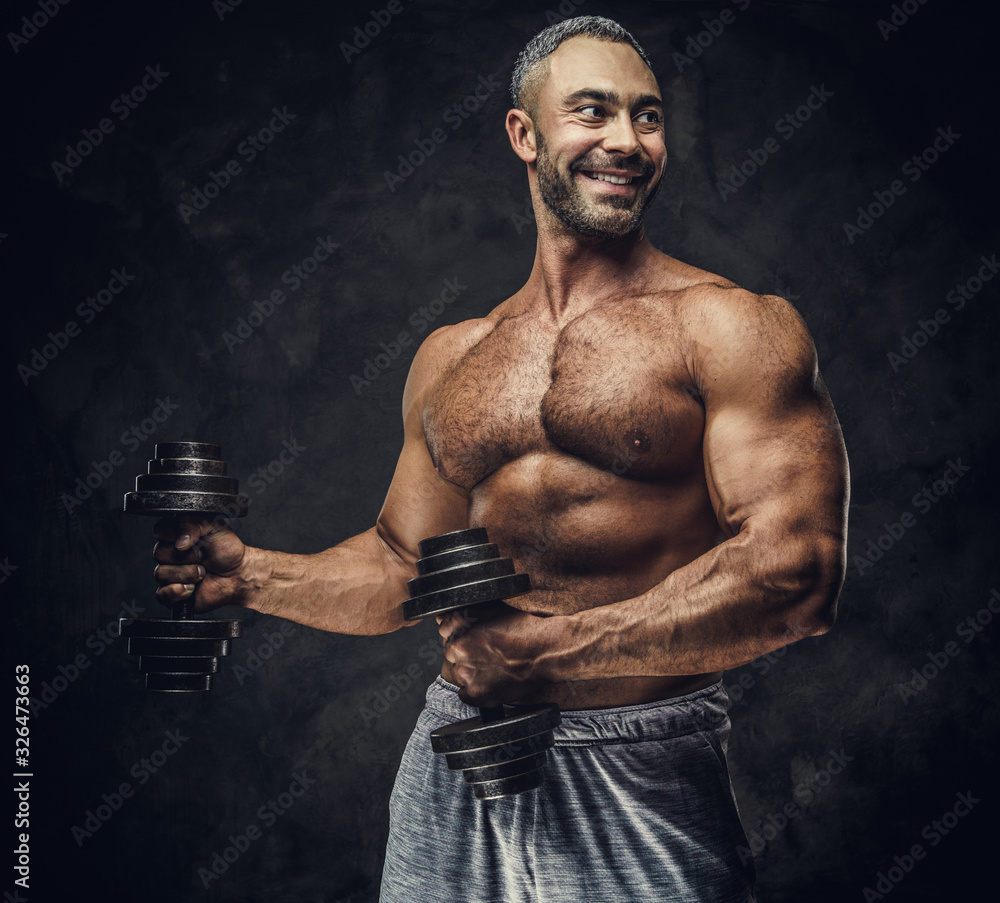 Strong, adult, fit muscular caucasian man coach posing for a photoshoot in a dark studio under the spotlight wearing grey sportswear, showing his muscles and putting up a dumbbells looking joyful
