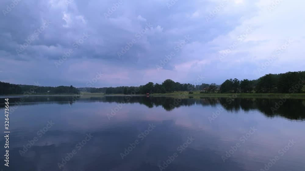Gorgeous view of sunset on calm summer evening. Lake shore with green trees and plants reflecting in crystal clean mirror water surface. Sky covered with heavy thunder clouds. Gorgeous view of sunset on calm summer evening. Lake shore with green trees and plants reflecting in crystal clean mirror water surface. Sky covered with heavy thunder clouds.