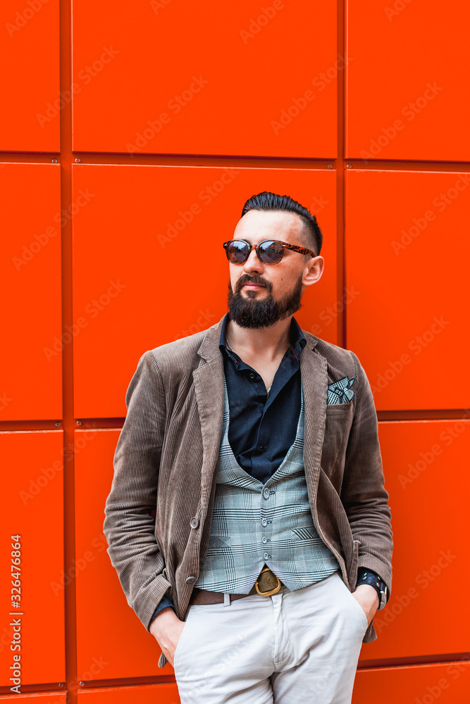 Young confident hipster man with beard in glasses posing on lush lava color background. Outdoor. Cute man wearing a vest, black shirt and light trousers. Man keeps hands in pockets and looking forward Young confident hipster man with beard in glasses posing on lush lava color background. Outdoor. Cute man wearing a vest, black shirt and light trousers. Man keeps hands in pockets and looking forward