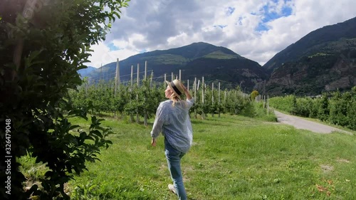 Young caucasian woman checking harvest in orchard walking near green trees row, hipster girl having tour to rural aric and agriculture on field photo