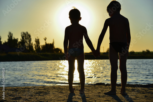 Two brothers are standing on the banks of the river and holding hands against the setting sun. photo