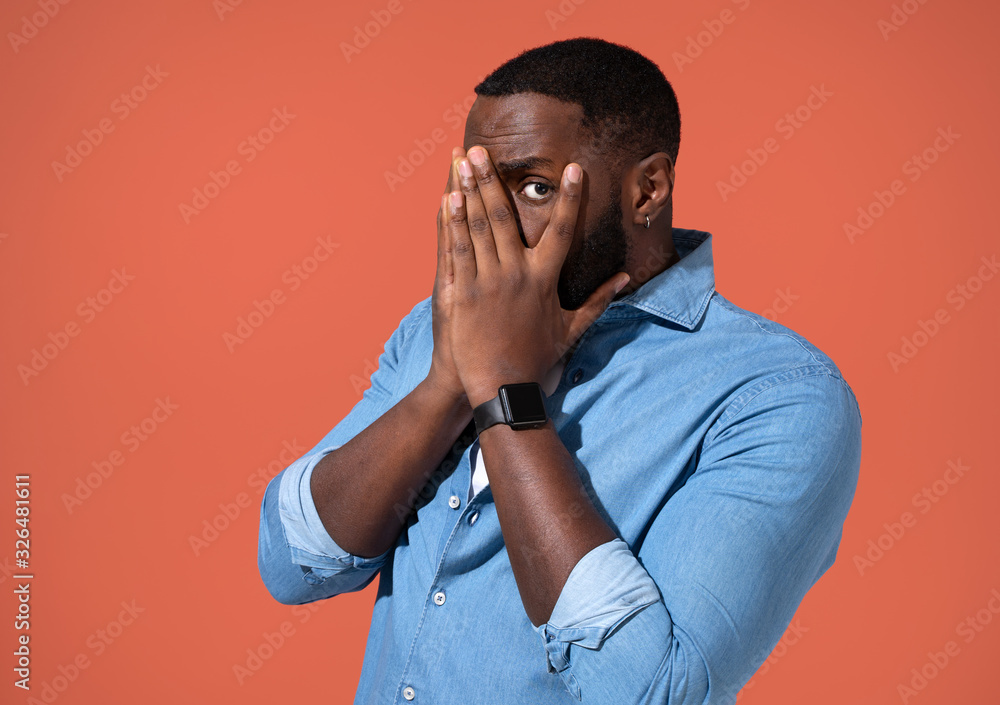 Frightened man peeks through fingers, covers face with both hands. Photo of african man in casual outfit on coral background. Frightened man peeks through fingers, covers face with both hands. Photo of african man in casual outfit on coral background.
