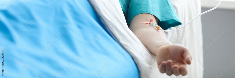 Fototapeta premium Fight for life concept. Hand of a young man with a dropper in ward of medical hospital. Lies after car crash in bed alone vegetable.