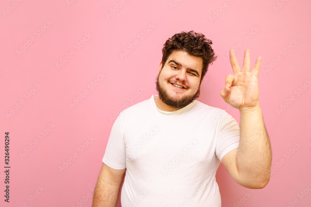 Fototapeta premium Smiling fat guy in white t-shirt shows thumbs up gesture, looks into camera and smiles. Cheerful young man with overweight and beard shows all right gesture on a pink background. Isolated.Copy space