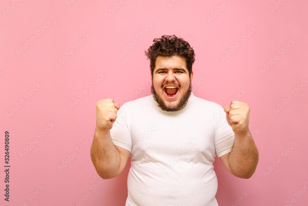 Portrait of a joyful guy in a white T-shirt and overweight rejoices the victory of his beloved team on a pink background. Happy fat cheerful man with beard raised his hands up for joy. Copy space Portrait of a joyful guy in a white T-shirt and overweight rejoices the victory of his beloved team on a pink background. Happy fat cheerful man with beard raised his hands up for joy. Copy space