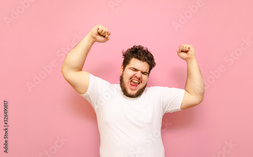 Happy fat young man shouting with joy with his hands raised on a pink background. Joyful overweight guy and white T-shirt, emotionally happy to win. Winning concept. photo